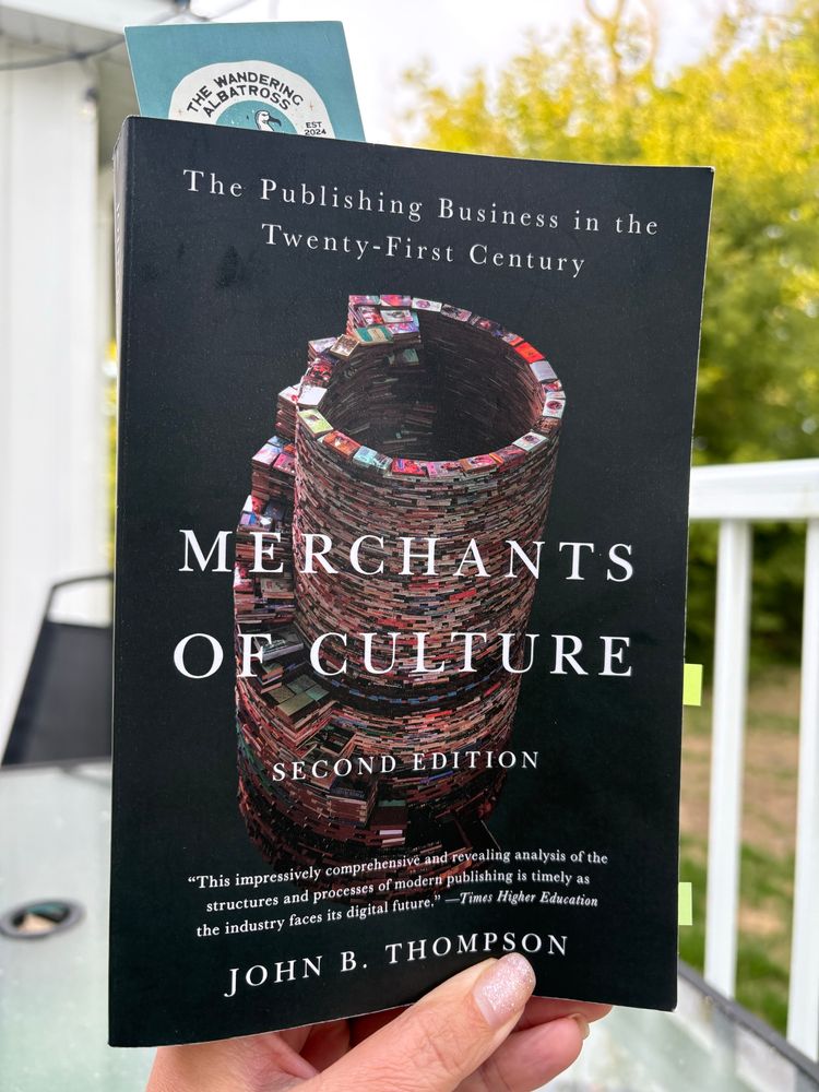 A hand holding a copy of the book Merchants of Culture by John B Thompson. The cover of the book shows a tower of books on a black background. In the background of the photo is a white deck railing and a green tree.