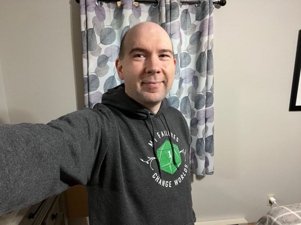 A white dude in his 40s, Calvin, stands modeling a charcoal grey hoodie with a 20-sided die on the front. The die has rolled a 1. Around the die are the words “my failures change worlds”