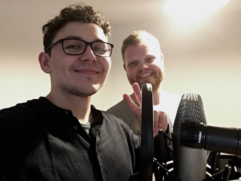 Selfie of Jake and me smiling. There are two microphones set up in front of us (SM57, and a large diaphragm condenser, not sure the make), with a pop filter attached. 