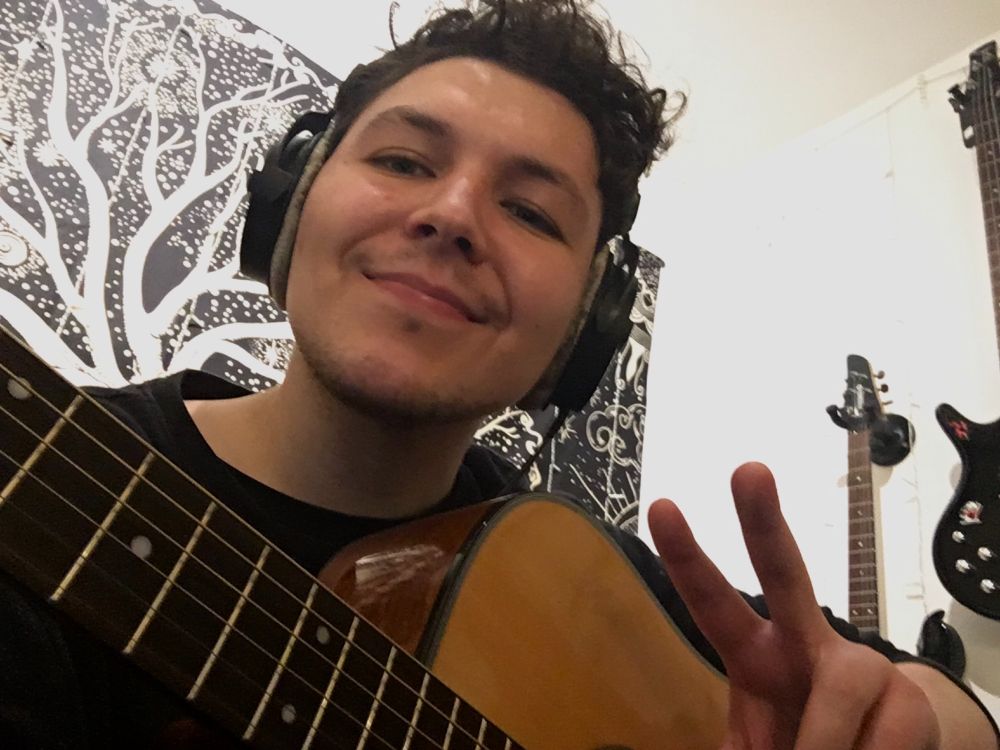 Selfie of me smiling, holding an acoustic guitar, throwing a peace sign. There is a guitar and bass hanging on a wall behind me.