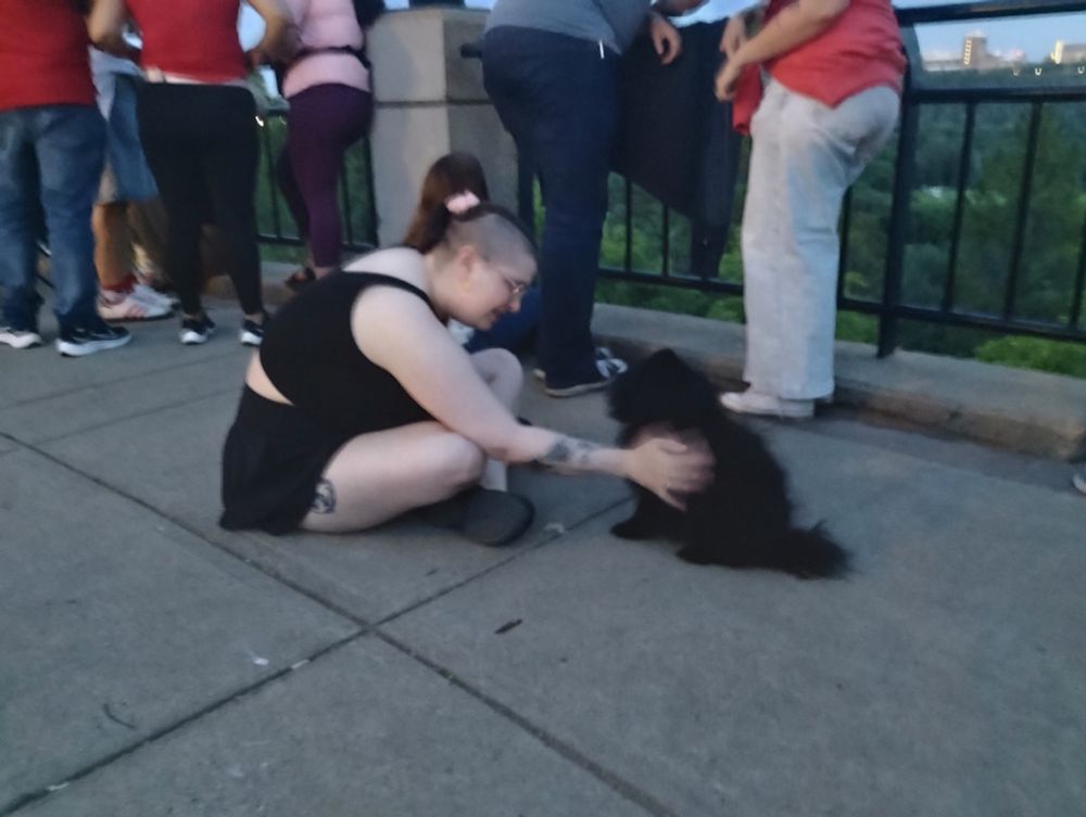 A blurry photo of Nicole sitting cross legged on the ground petting a black Pomeranian 