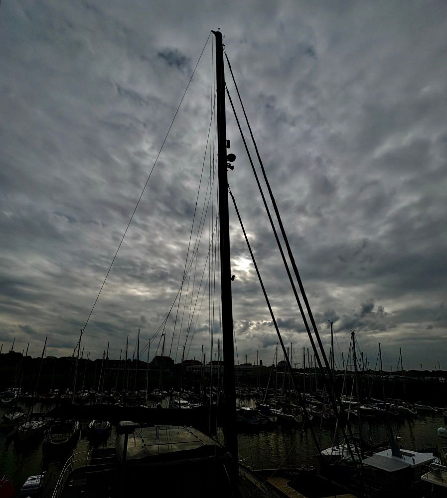 Vor einem bewölkten Himmel mit einer nicht völlig sichtbaren Sonne erhebt sich riesengroß ein schwarzer Mast mit seinen Verstrebungen umliegende Yachten sind nur schemenhaft klein zuerkennen, sehr schmal und klein ihre Masten 