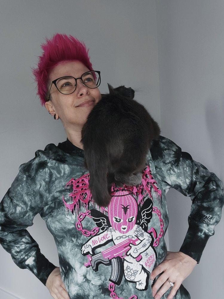 A white human with pale skin, short pink hair, and black framed glasses makes a silly pose, smirking with hands-on-hips, with a gray cat on their shoulder.