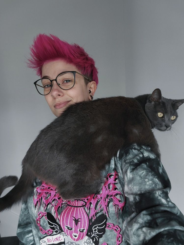 A white human with pale skin, short pink hair, and black framed glasses smiles at the camera with a gray cat on their shoulder.