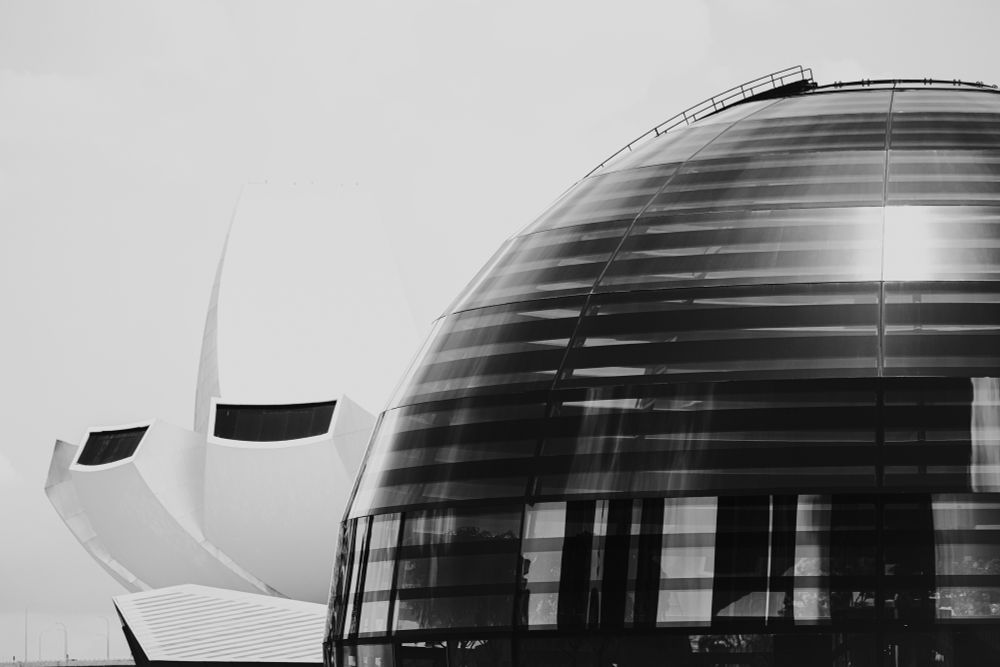 A black-and-white photograph featuring two prominent modern buildings. On the right side, there is a dome with horizontally segmented glass panels that reflect ambient light and partially reveal an interior framework. To the left, a bright white structure with multiple curved segments appears, evoking the look of layered petals or sculptural waves. The sky behind them is light and featureless, accentuating the contrast between the dark glass and the smooth white surface. 