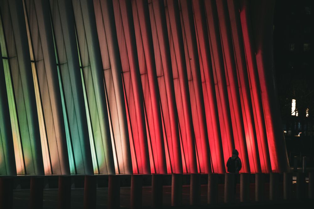 A person in a hooded jacket with a backpack walks past a modern architectural structure at night. The structure features tall, vertical beams illuminated by a gradient of green, yellow, and red lighting. The beams are arranged in a slightly slanted formation, creating a sense of depth and movement. The figure appears small in comparison to the towering structure, emphasizing a contrast between human scale and urban design. In the background, faint lights from buildings and street lamps are visible, adding context to the nighttime city scene.
