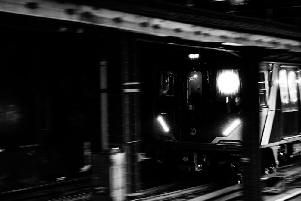 A black-and-white photograph of a subway train speeding through an underground station. The image is taken from a slightly oblique angle, capturing the train’s headlights glowing in the dark tunnel. The platform above partially obscures the train, adding a sense of movement and urgency. The lights inside the train reveal the faint outline of the driver, while the exterior appears blurred, emphasizing its rapid motion.