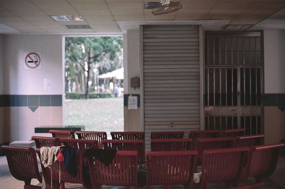 A film photograph of a scene at the void deck of the HBD block in Singapore. A series of red metal chairs are laid out in partial concentric circles with some clothes being hung over the back of the last row of chairs. There is some greenery in the background