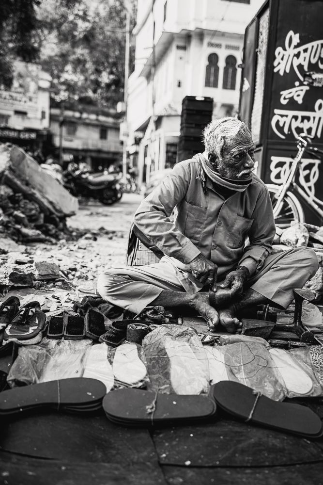 A black and white image of a cobbler on the streets of India. The cobbler is sitting down on a makeshift plastic mat and is surrounded by various shoes and shoe components.

His face has a prominent mustache and he looking away from the perspective of the camera 