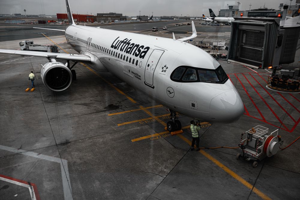 A Lufthansa Airbus A320neo is stationed at the gate at an airport,just after arrival. The aircraft is white, with the airline's logo and name prominently displayed along the fuselage. Ground crew members in high-visibility vests are attending to aircraft, with one person standing near the nose wheel and another by the left wing. The aircraft's jet engine is visible on the left, and a jet bridge is retracted in the background. Several other planes and airport service vehicles are scattered across the tarmac under an overcast sky.