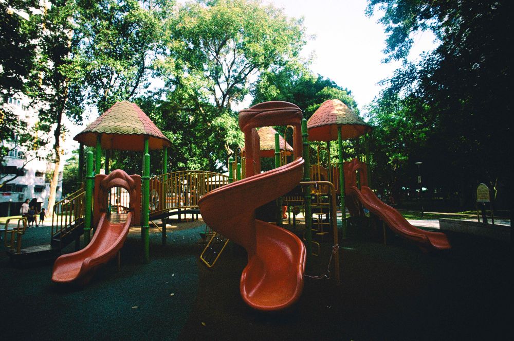 Film photograph of a playground in Singapore. There are 3 red slides in focus backed by stairs and small hut-shaped play structures. There are HDB blocks and trees in the background