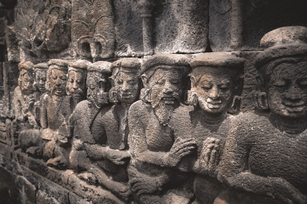A close-up view of an ancient stone relief depicting a row of human figures carved into the rock. The figures, adorned with headgear and earrings, appear to be seated in a meditative or devotional posture, their hands clasped together. The texture of the stone is rough and weathered, with visible cracks and erosion. 