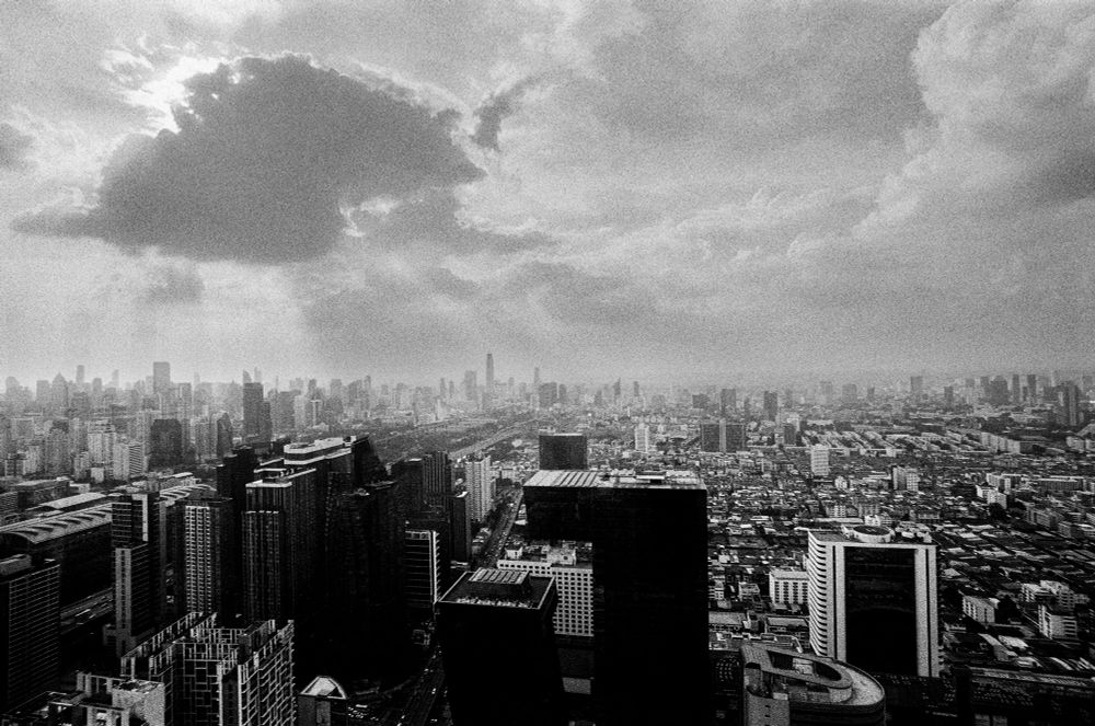 A black-and-white image shows a high vantage view of a sprawling city. Multiple tall buildings dominate the foreground, extending outward in a dense arrangement toward the horizon. A prominent cloud formation appears overhead, partially blocking sunlight, resulting in varying degrees of brightness and shadow across the scene. The cityscape includes layered blocks of structures, with roads and smaller buildings receding into the distance.