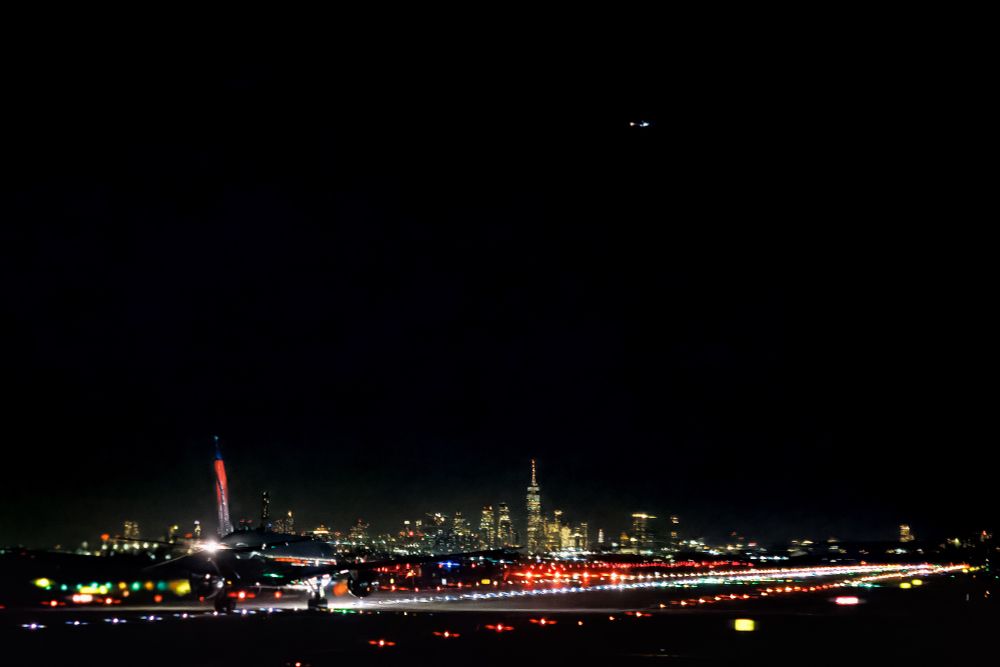 A nighttime photograph of JFK airport in NYC. A plane is on on the runway accelerating to take off. The headlights of the plane illuminate the runway.

The skyline of NYC is in the background