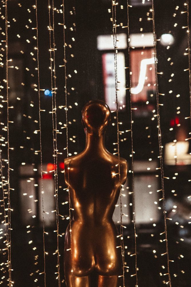 A golden mannequin torso stands facing away from the camera, surrounded by hanging string lights. The lights create a warm, glowing atmosphere, with small, twinkling bulbs suspended vertically in the foreground. In the background, a blurred cityscape with neon signs and reflections in a glass window adds depth. The mannequin’s metallic surface subtly reflects the surrounding lights, enhancing its golden hue.
