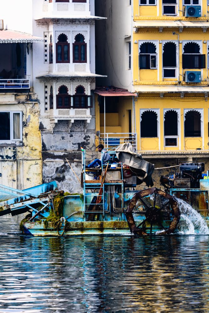 A mechanical water-cleaning boat sits in a river against a backdrop of old, brightly painted buildings. The boat features a rusty paddle wheel and machinery covered in moss, with a man operating the controls. The architecture showcases ornate, arched windows and weathered walls, blending traditional design with signs of modern decay. The water below reflects the vivid colors and textures