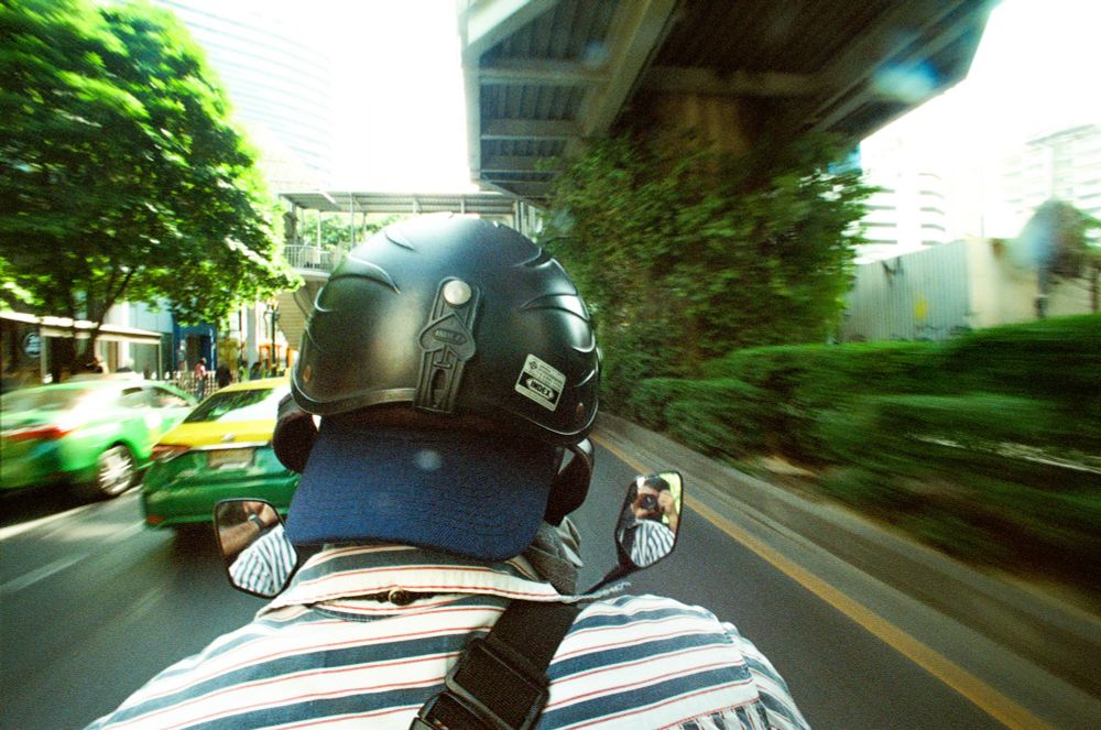 An individual wearing a black protective helmet and a striped collared shirt is seen from behind on a moving vehicle in an urban setting. Two rearview mirrors flank the helmet, reflecting portions of the rider and surroundings. The photograph captures motion blur of passing green and yellow taxis and tall city buildings in the background. Overhead structures cast shadows across the roadway. The rider’s camera strap is visible on the shoulder, indicating a camera is in use. Lush greenery appears along the right side, contrasting with the roadway. The image is composed with the subject centrally framed and the cityscape receding around them.