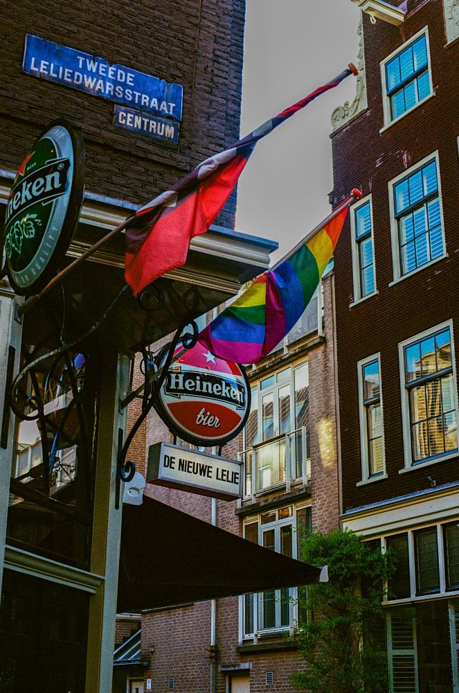 A lively urban street scene in Amsterdam. The image features the sign for "De Nieuwe Lelie" with a Heineken beer logo prominently displayed. Above the sign are two flags, one a solid red and the other a rainbow Pride flag. The corner of the building includes a blue street sign reading "Tweede Leliedwarsstraat Centrum." Surrounding buildings exhibit traditional Dutch architecture with brick facades and large windows.