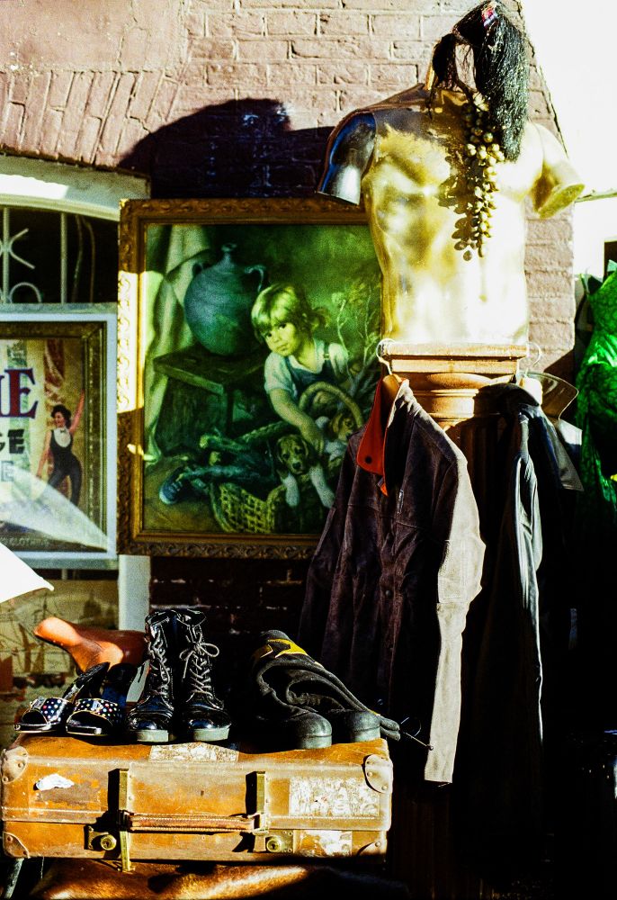 A cluttered display outdoors includes a headless, golden mannequin wearing a necklace of grapes, standing on a pedestal. Next to it hangs a brown coat, and on a worn suitcase below are various shoes, including black lace-up boots and studded high heels. In the background, a framed painting of a child sitting near a basket and a vintage circus poster add to the eclectic scene.