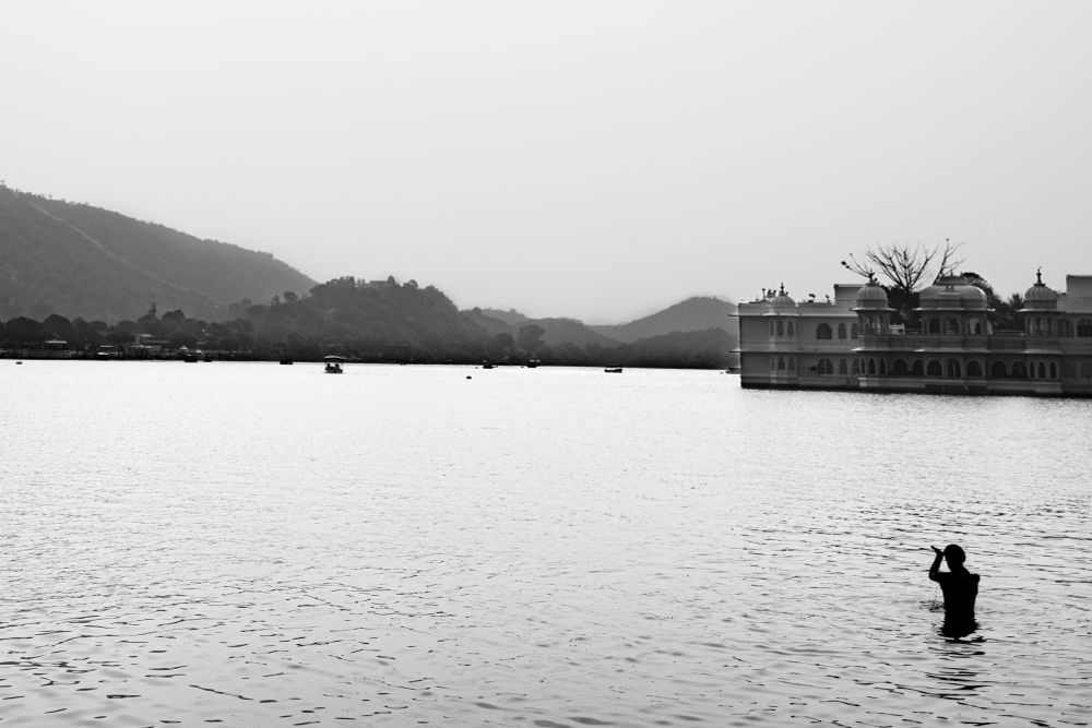 A black and white photograph of a Serene Lake with faint ripples. In the foreground, a silhouette figure stands waist deep in water, seemingly raising their arms.To the right, an ornate palace rests on the lake surface, showcasing, dorms and arches. The background features distant hills, feeding into a hazy horizon.
