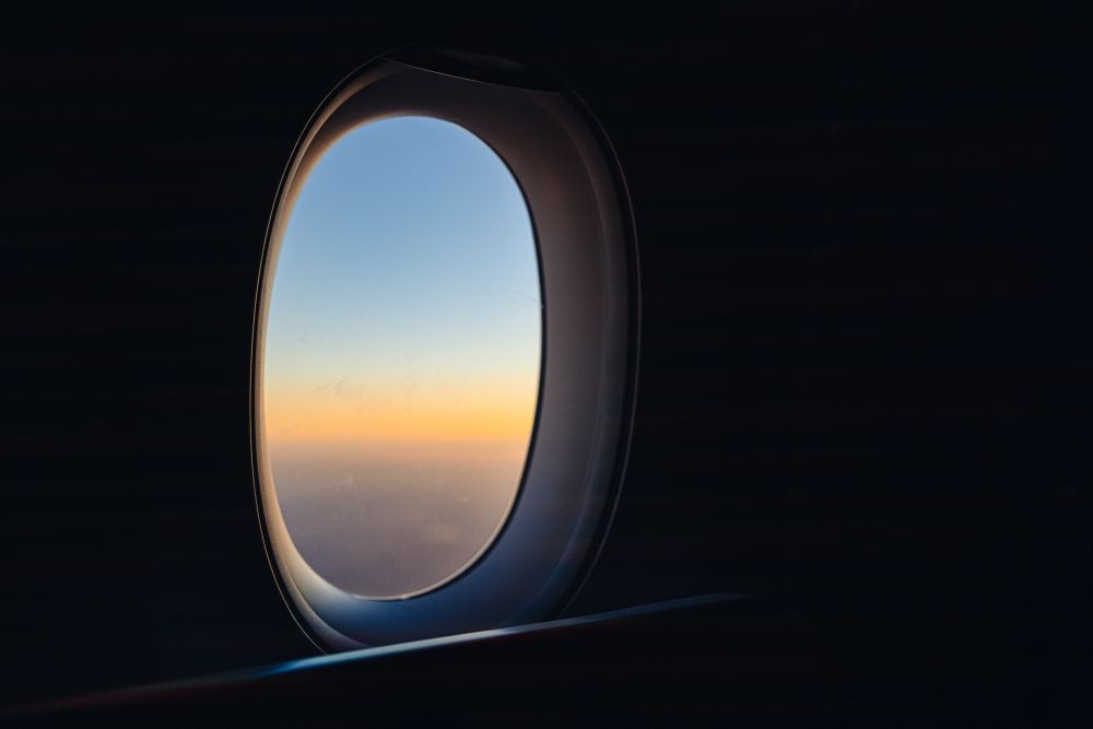 The image captures the view through an airplane window during flight, showcasing a gradient of colors in the sky transitioning from a deep orange near the horizon to pale blue above. The window frame and the dim interior of the aircraft form a stark contrast against the vividness of the sky outside. No other objects or features are visible beyond the horizon line.