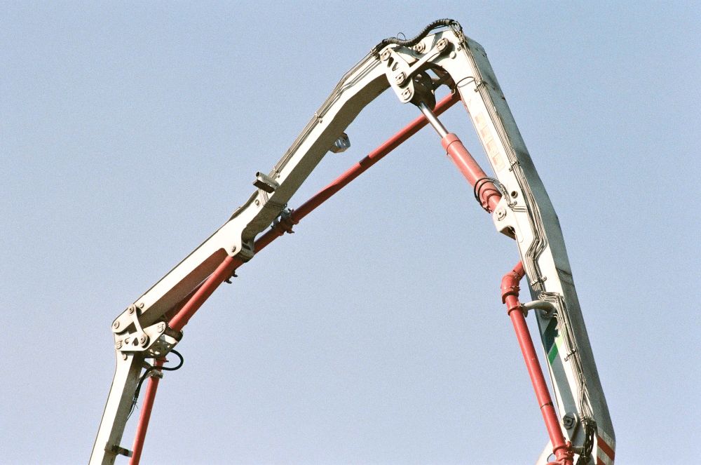 A film photograph of an mechanical articulated arm from construction equipment. The photograph is framed in such a way that the arm follows the shape of a parabola. The background is a light blue sky without any clouds