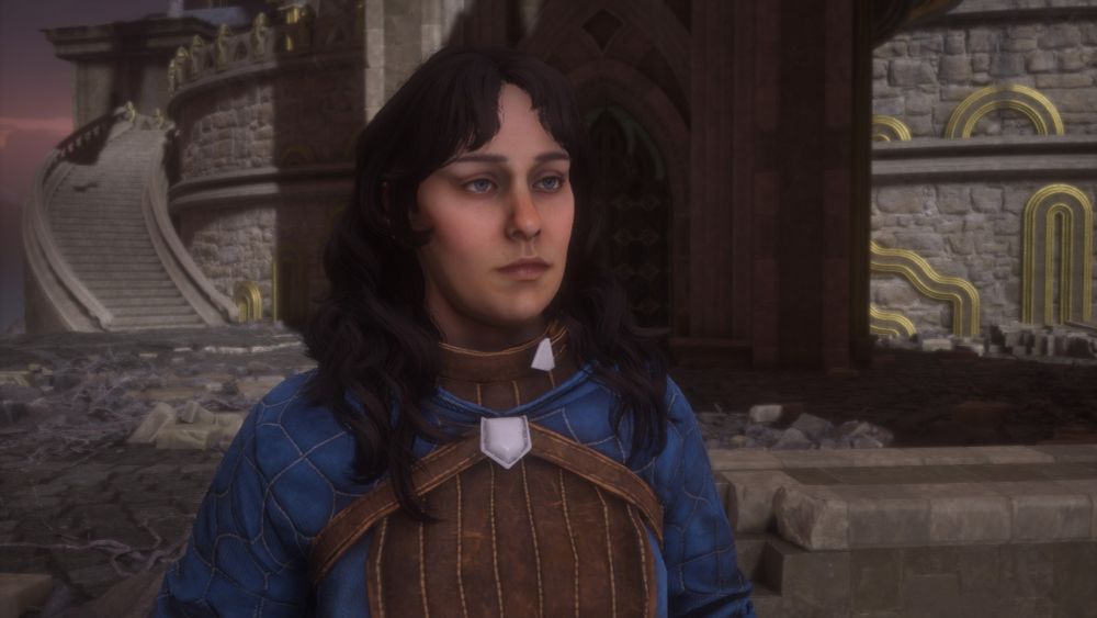 A woman with long, curly brown hair wearing a blue and brown shirt stands in front of a wooden doorway and stone staircase.