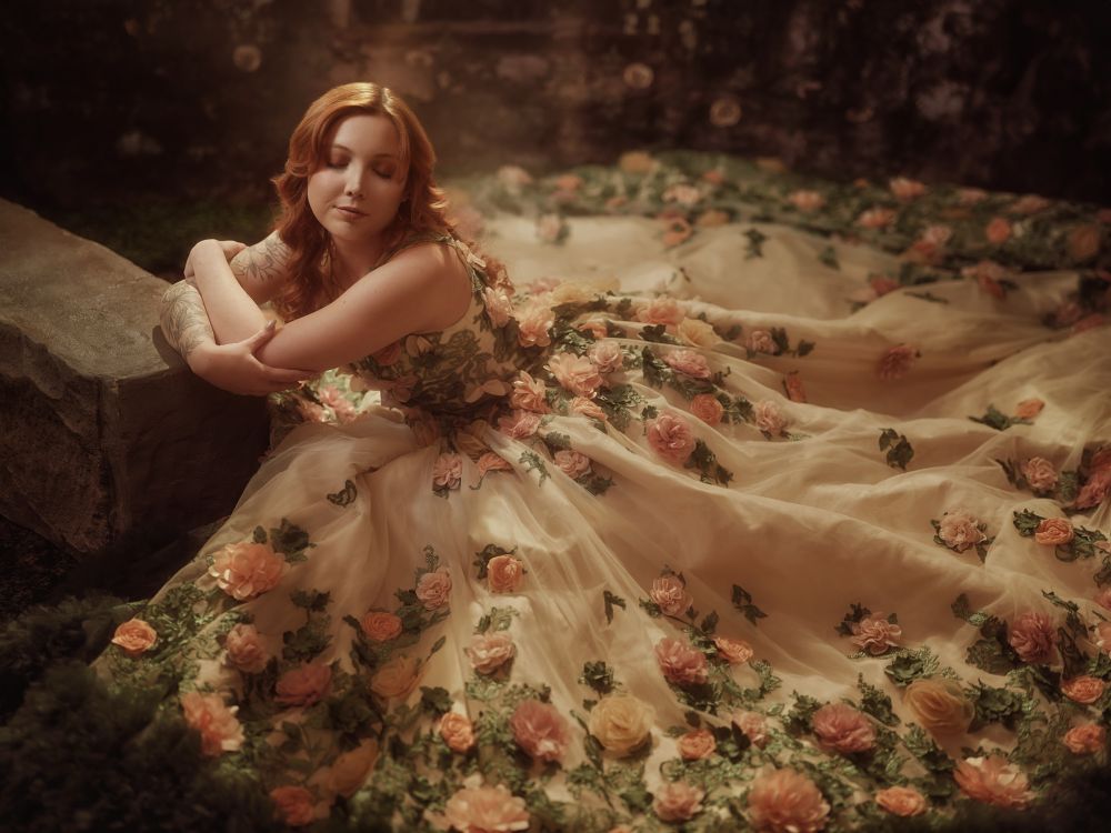 Woman in gown with flowers sits on the ground in warm glowing light 