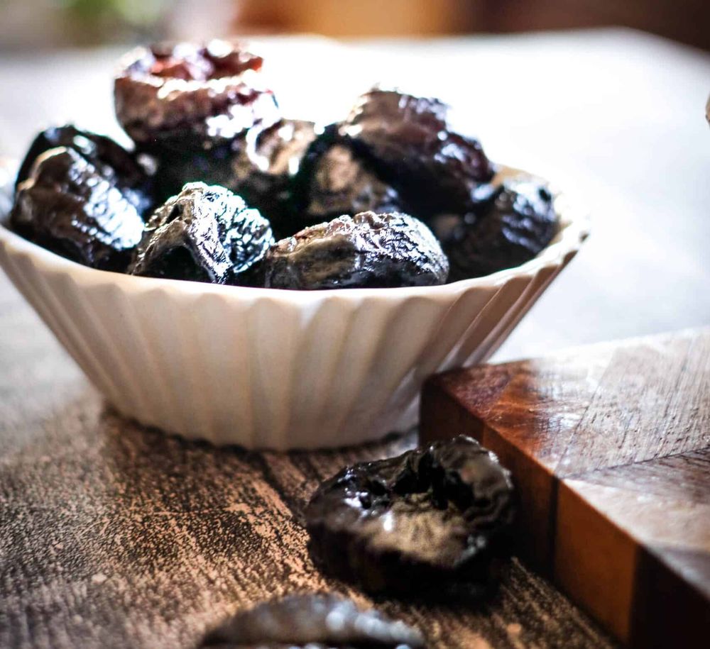 prunes in a fluted ceramic cup, prunes prunes prunes they are dark, the cup is white, they are on a wood table, prunes!!! oh god, what are we all doing here?