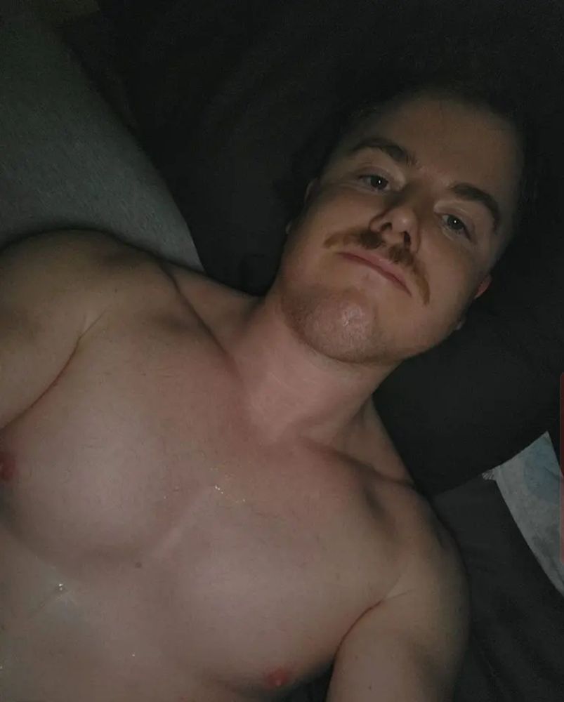 Andy is laying in bed shirtless with a white fluid in ropes on his chest. His facial expression is one of satisfaction.