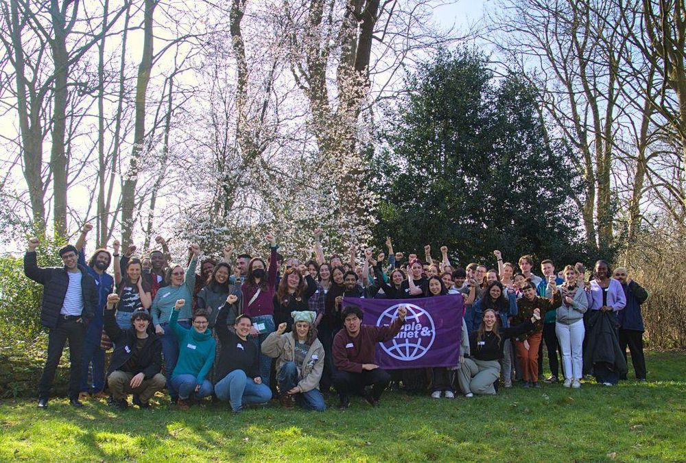 Photo of a crowd of about 40 people, most with fists in the air and big smiles standing outside on grass with trees behind on a sunny day. The crowd is a diverse group of young people.