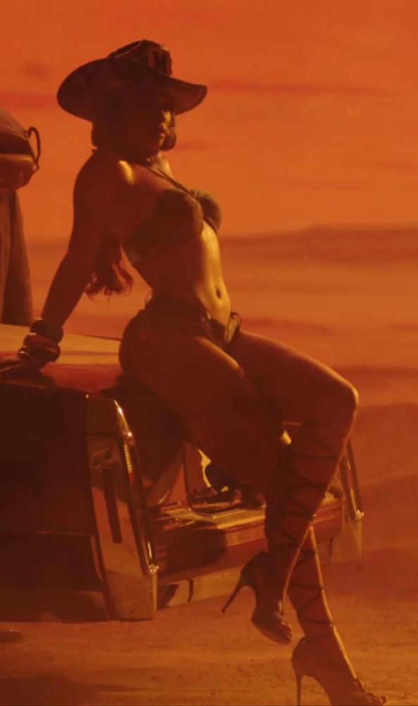 Rapper Megan Thee Stallion sitting on the back trunk of an old car with her legs crossed at the thighs. She wears a cowboy hat, daisy dukes and a small bikini to, lace up heels on her feet. 
