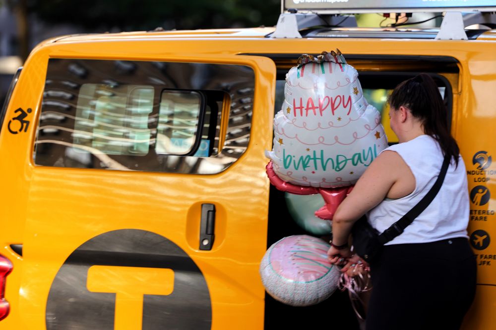 A woman loads birthday balloons into a yellow NYC taxi.