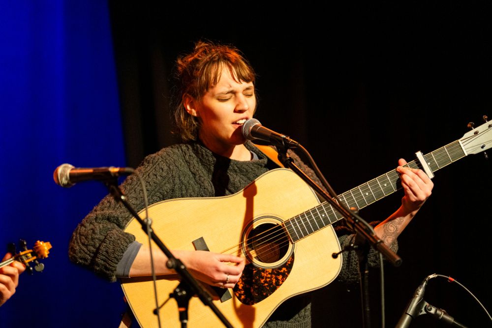 A brown haired individual wearing green pants, Helen Kuhar, plays a guitar and sings into a microphone. 