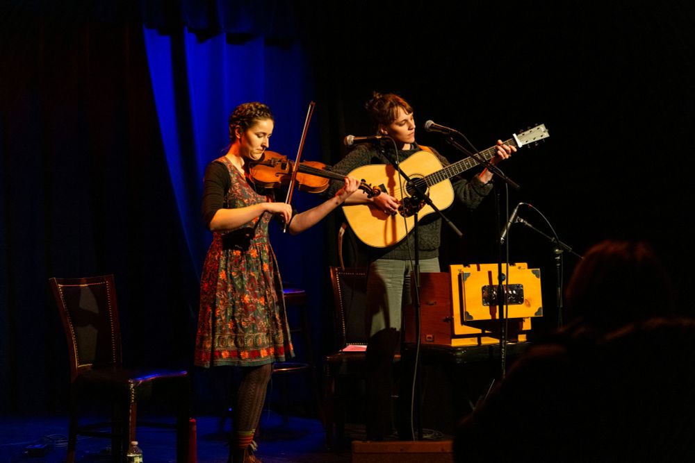 A brown haired individual, Rose Jackson, wearing a floral dress stands on the left on a stage in front of a blue backdrop, playing a fiddle. A brown haired individual wearing a gray sweater and green pants, Helen Kuhar, stands to the right playing a guitar.