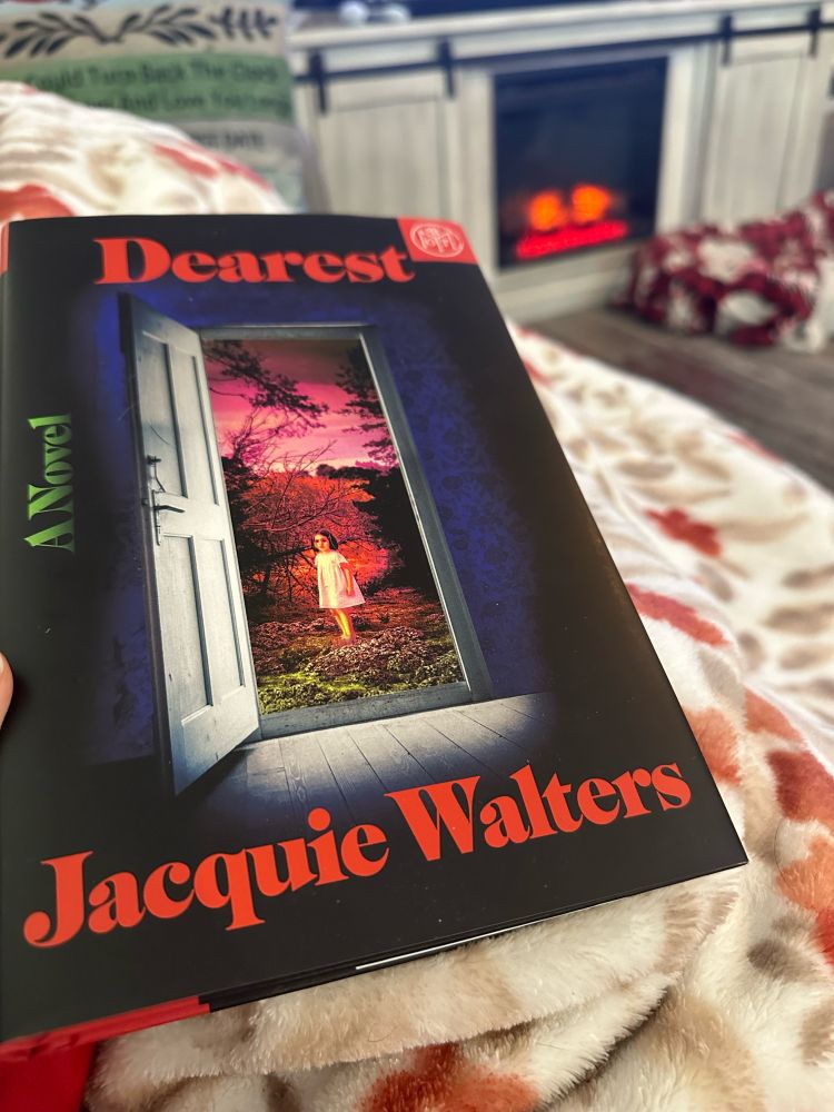 Book cover of Dearest, by Jacquie Walters, showing an open door to a pink sky and a little girl standing in some brush. Book is on a blanket with a fake fire burning in the background. 