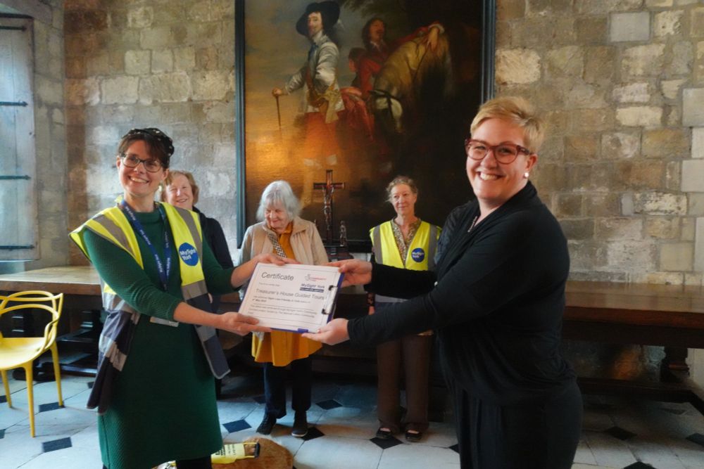 Vicky from MySight York is presenting a certificate to Rebecca from Treasurer's House. A group of our members and volunteers are in the background, standing in front of a huge painting.