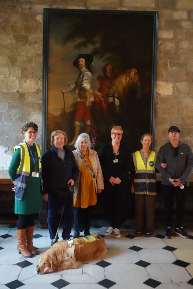 A group of 6 people, including staff, members and volunteers of MySight York and a staff member of Treasurer's House are standing in front of a huge painting. There is a relaxed Guide Dog snoozing on the floor.