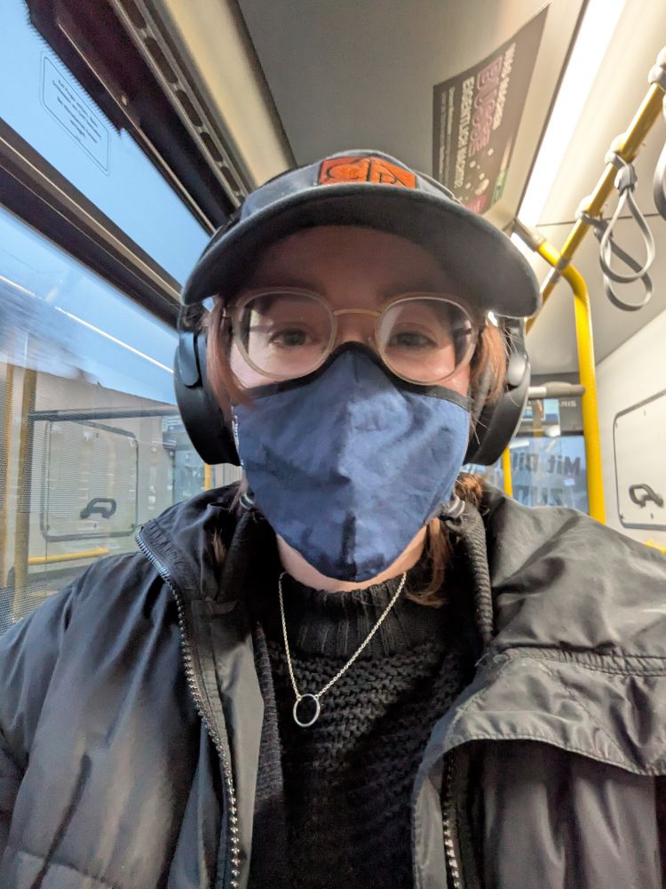 A selfie of Hunter, a nonbinary person with white skin, in a bus wearing a critical role baseball hat, glasses, noise cancelling headphones and a dark blue face mask. They're wearing a black knit sweater and a black winter coat that is zipped open over it.
