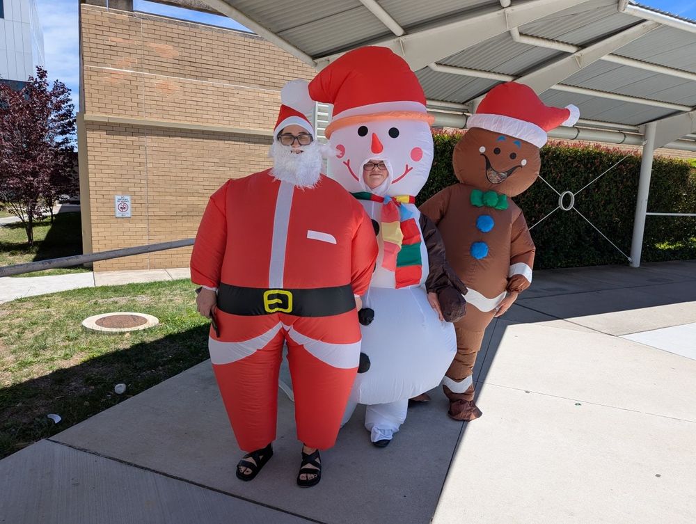 Three young women in inflatable Christmas suits (Santa, snowman, gingerbread man) under the narrow shade over the walkway outside Canberra Hospital. Outside of the shade it is very bright and sunny. Temp is 29.5 °C, so trad costumes are death.