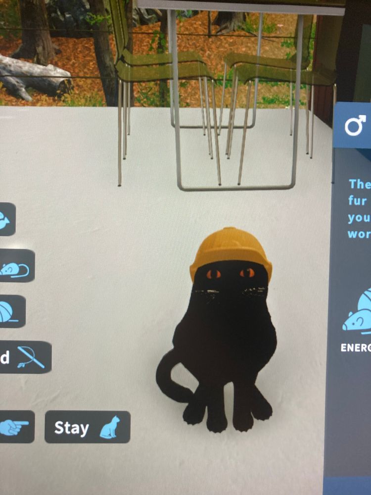 A video game screenshot of a black cat sitting on the ground. He looks more like a black blob. He wears a yellow hard hat on his head. 