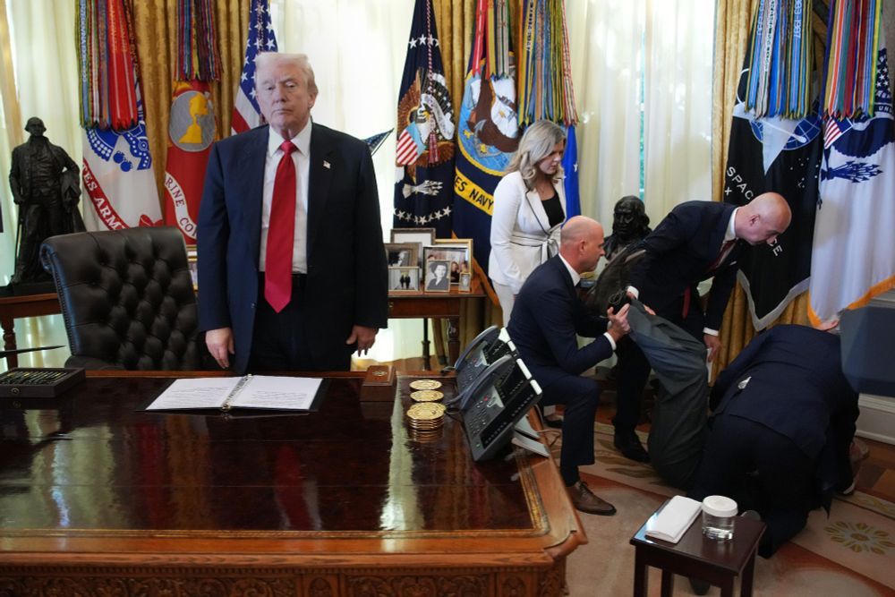 Donald Trump in the Oval Office,  standing perfectly still and expressionless, looking away as people cluster around someone who passed out.