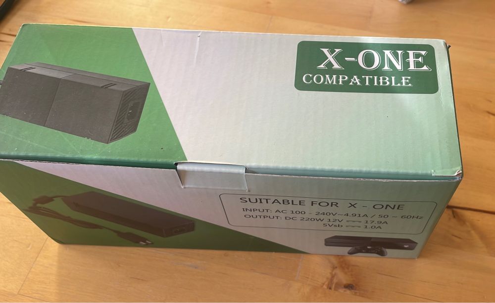 A box for a power brick suitable for Xbox one. The top of the box shows a picture of the brick on green, and a green box with rounded edges and the words “X-One Compatible” in a decorative font that is all caps, serif and has a built in 3D edge.