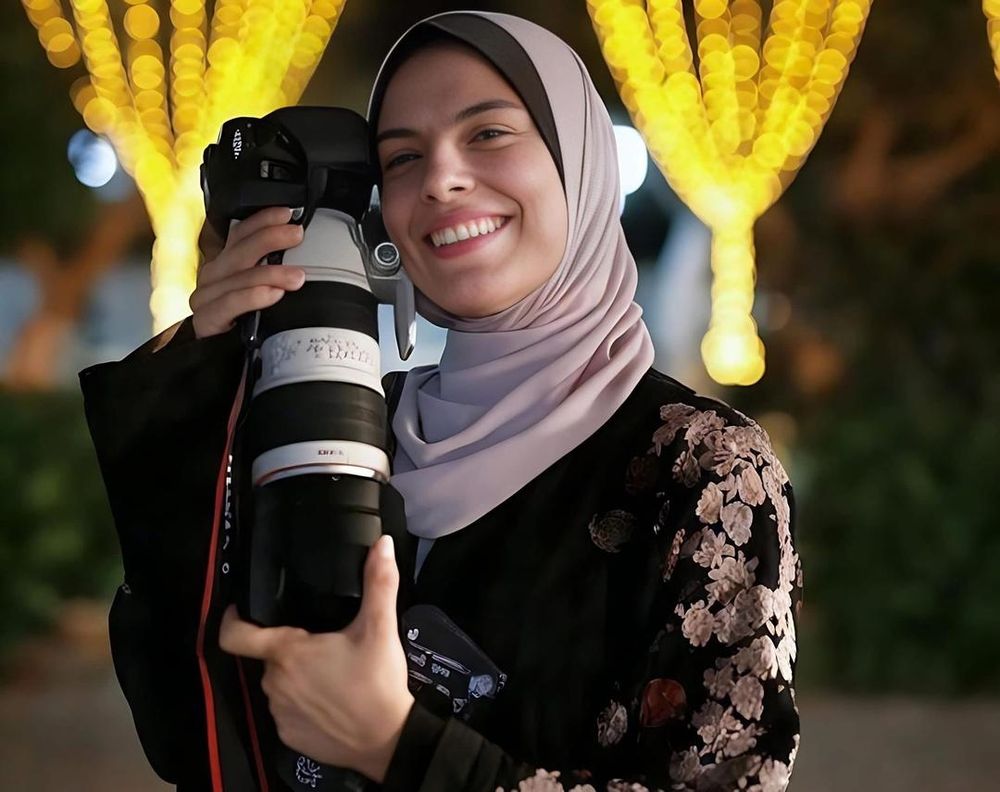 A higher resolution AI upscaled version of the same image. Her cheeks are rounder and more flushed with pink tones. Facial blemishes are smoothed out. The text on the camera lens is replaced with gibberish. The left side of her face that was previously cast in shadow and hard to see, is now is fully rendered and visible.
A subtle shadows and facial characteristics (like a shadow where her forehead meets her hijab) is just completely erased and smoothed out.