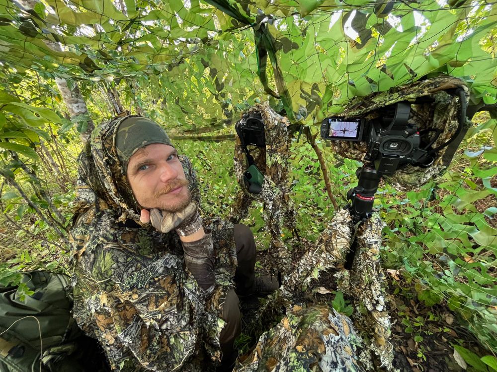 Mark O'Hara sits in a tree with camera equipment. Everything is fully dressed in camouflage. Mark looks happy to be there. 