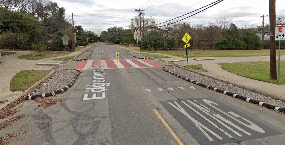 A photo of Northaven Trail in Dallas crossing Edgemere Road. The crossing has several safety features, besides a painted crosswalk and other visual elements. Bump outs from the curbs on either side of the crossing narrow the roadway and compel drivers to slow down and pay more attention. A stop-sign flexpost in the middle of the crossing does the same. An example of a good—by local standards—unsignalized intersection.