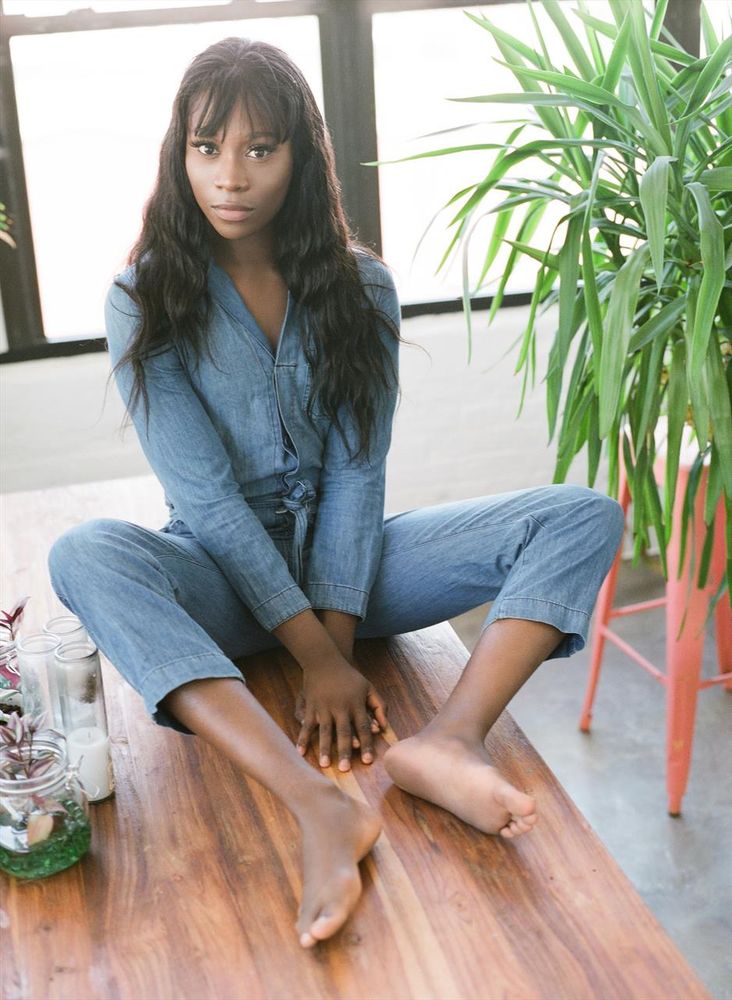 Vanniall sits on the floor barefoot wearing a matching denim shirt and pants. She is surrounded by plants and candles.