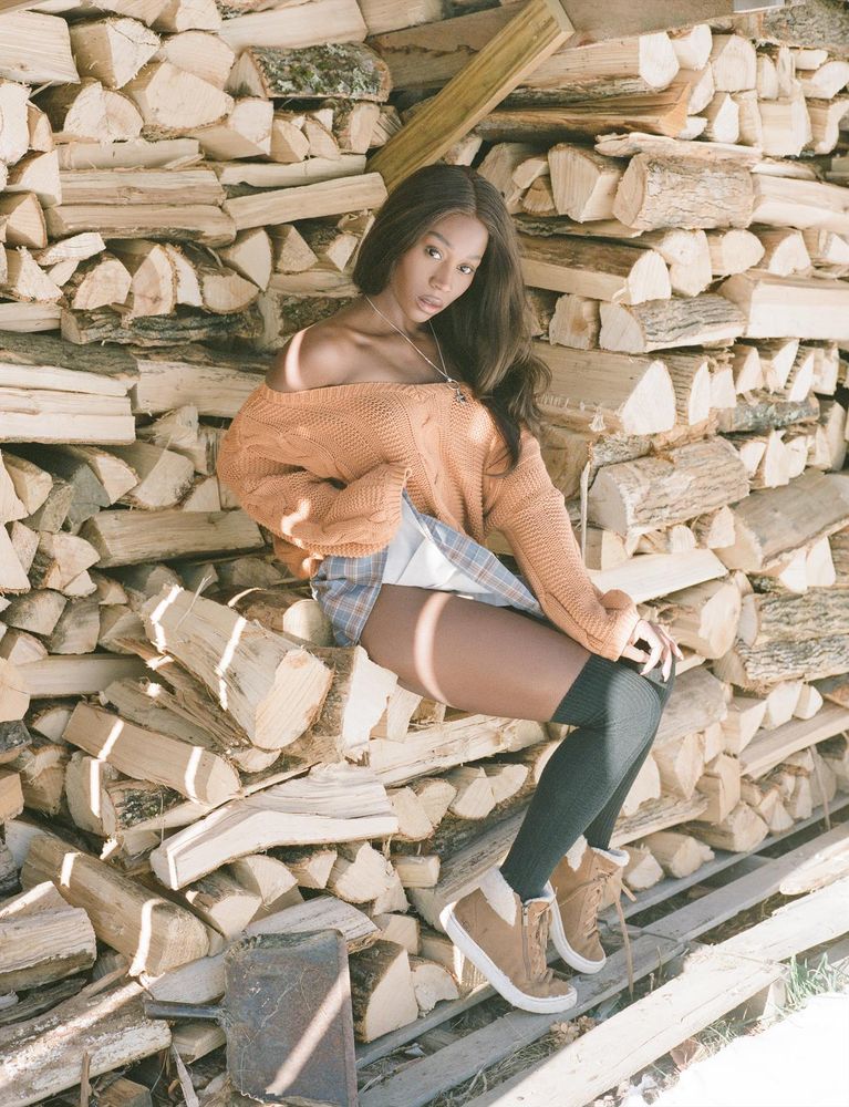 Vanniall sits on a big pile of chopped wood, wearing an orange sweater and plaid miniskirt. She has a smouldering look in her eyes and is lifting the hem of her skirt suggestively. 