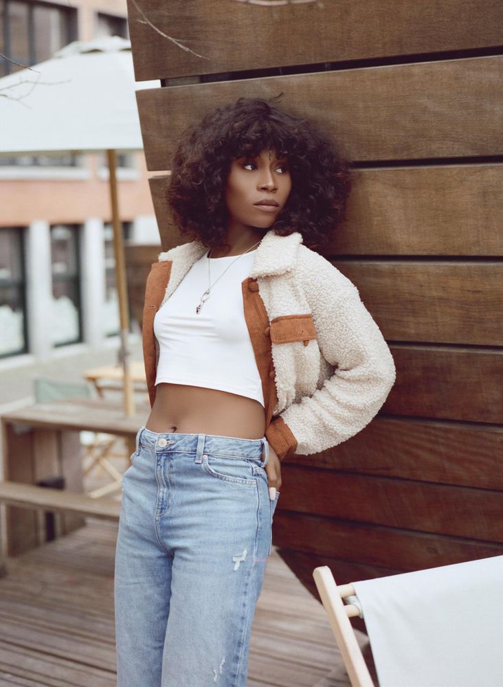 Vanniall leans on a curved wooden structure, wearing jeans, a cropped white shirt, and a shearling jacket. She is looking away from the camera.