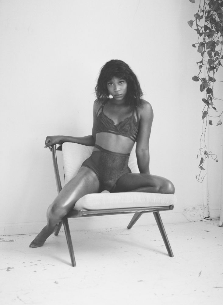 Vanniall poses on a chair wearing lingerie and making come-hither eye contact with the camera. The photo is black and white so the shape of her body and shine of her skin is accentuated.
